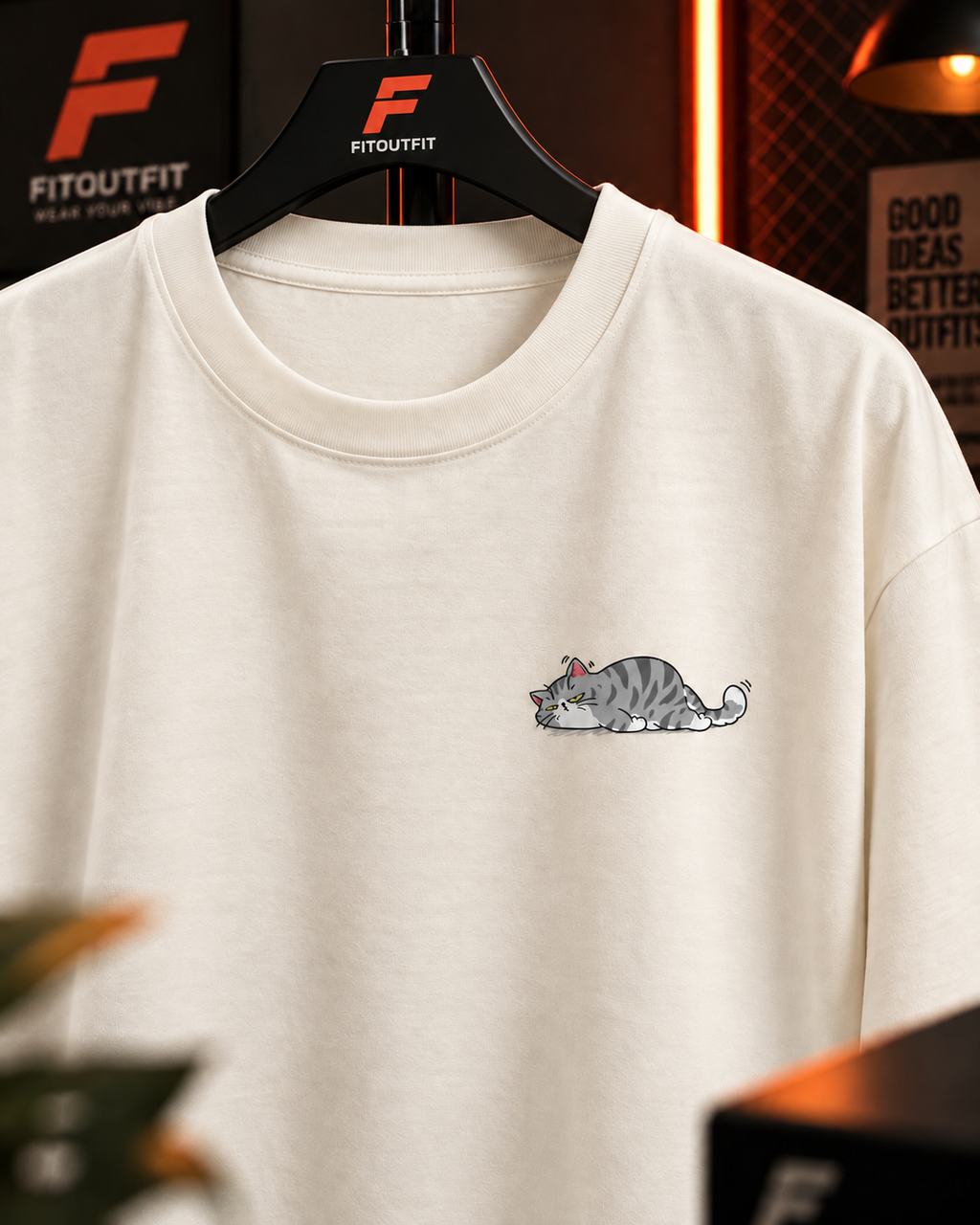 An off-white crewneck sweatshirt on a black hanger, featuring a small cartoon illustration of a sleeping gray tabby cat on the left chest. The hanger is branded with the red and black 'FITOUTFIT' logo, and the background shows a warm, dimly lit, stylish interior with blurred elements.