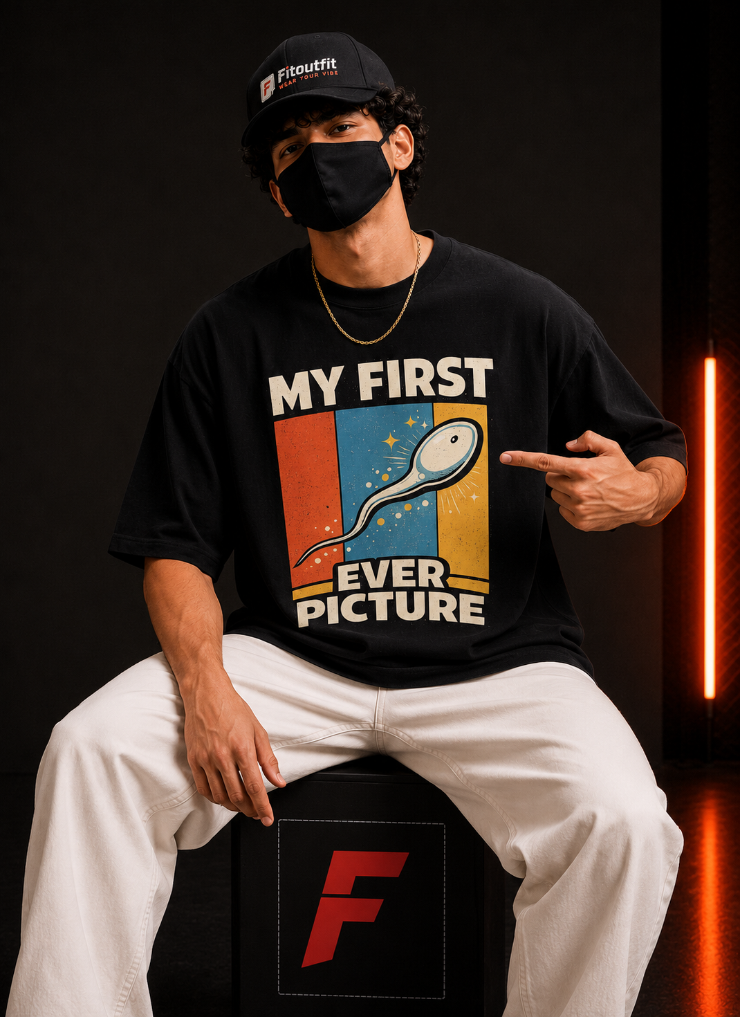 A man wearing a black graphic t-shirt with a retro-style sperm illustration and the text 'MY FIRST EVER PICTURE' sits on a black stool. The background is dark with a vertical orange light strip on the right.