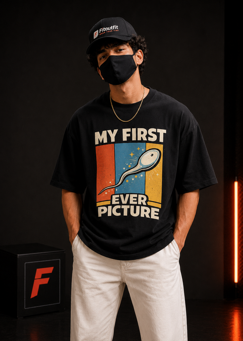 A man wearing a black graphic t-shirt with a retro-style sperm illustration and the text 'MY FIRST EVER PICTURE' in white. He is also wearing a black cap with a visible 'Fitoutfit' logo, white pants, and a black face mask, standing against a dark studio background with warm lighting.